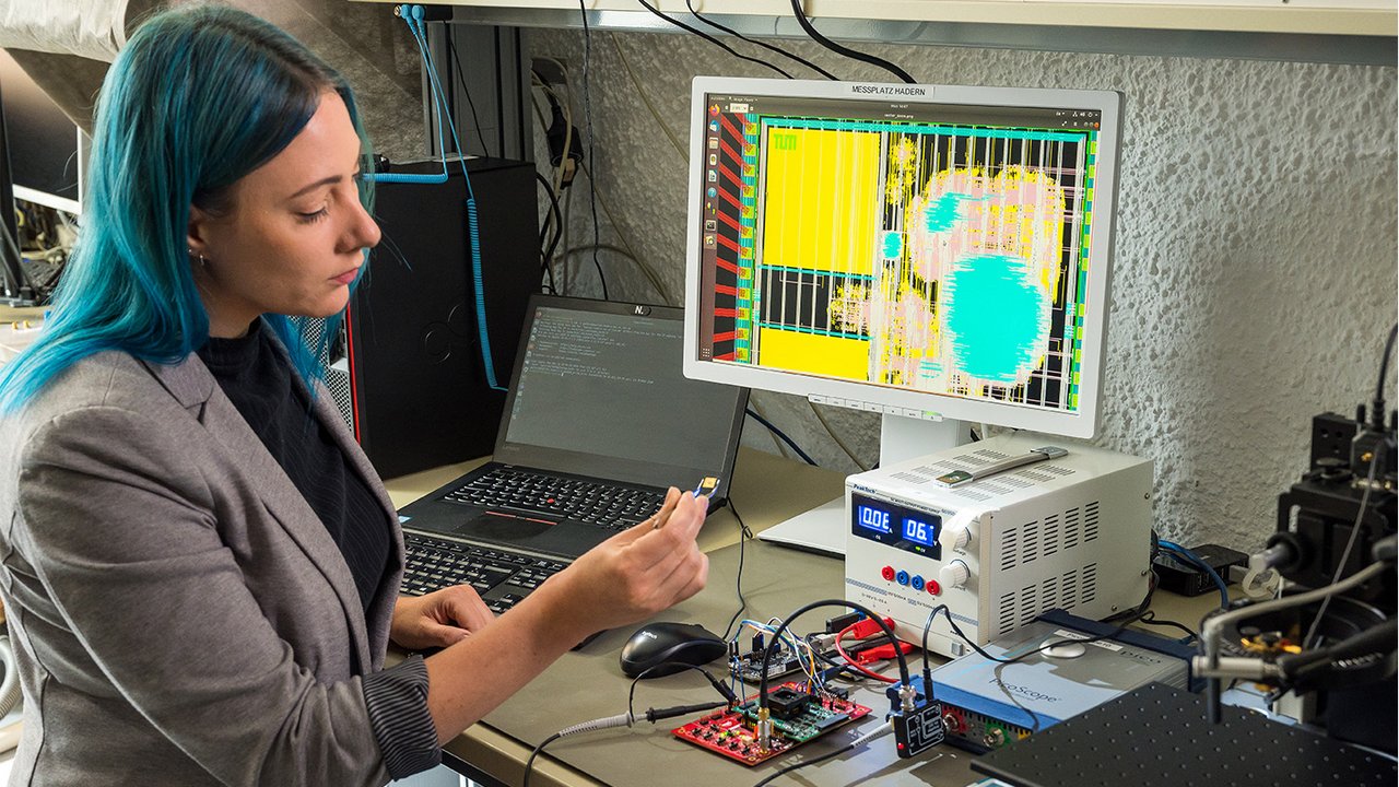 A scientist working with a computer chip in front of computers and monitors.