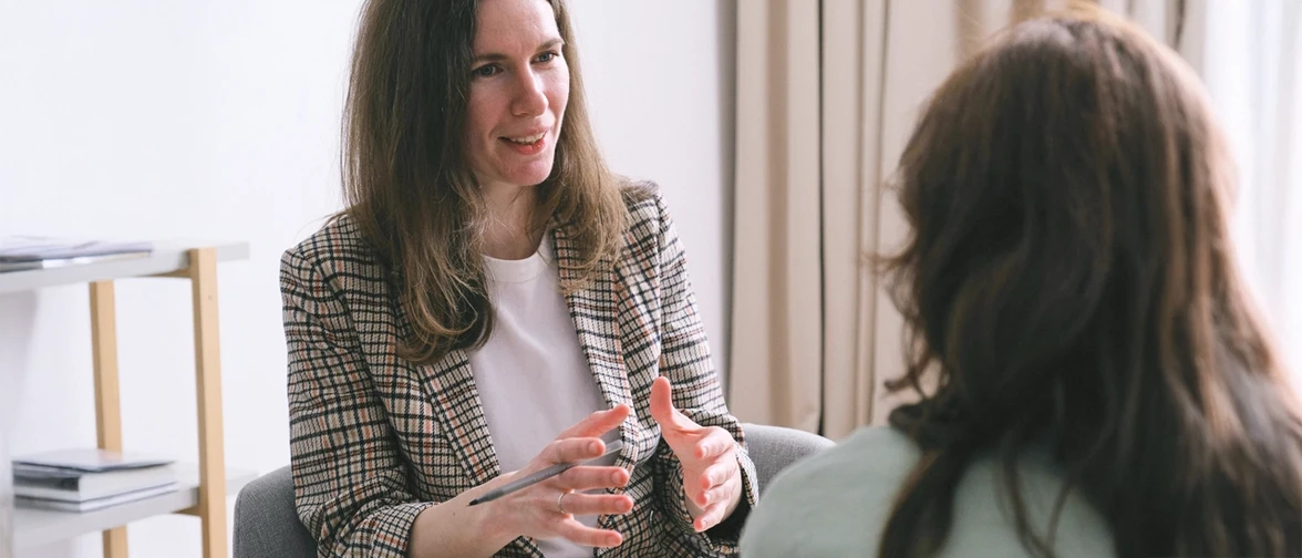 Two people sit facing each other in a bright room, engaged in conversation, with one gesturing expressively while holding a pen, suggesting a professional discussion or meeting.