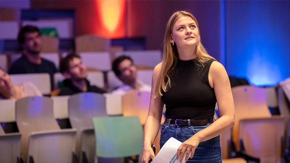 A woman stands in an auditorium holding papers, addressing or preparing to address a seated audience, with colorful stage lighting and rows of empty and occupied chairs around her.